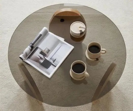 Round glass coffee table with two cups of coffee and a magazine on a light-colored floor.